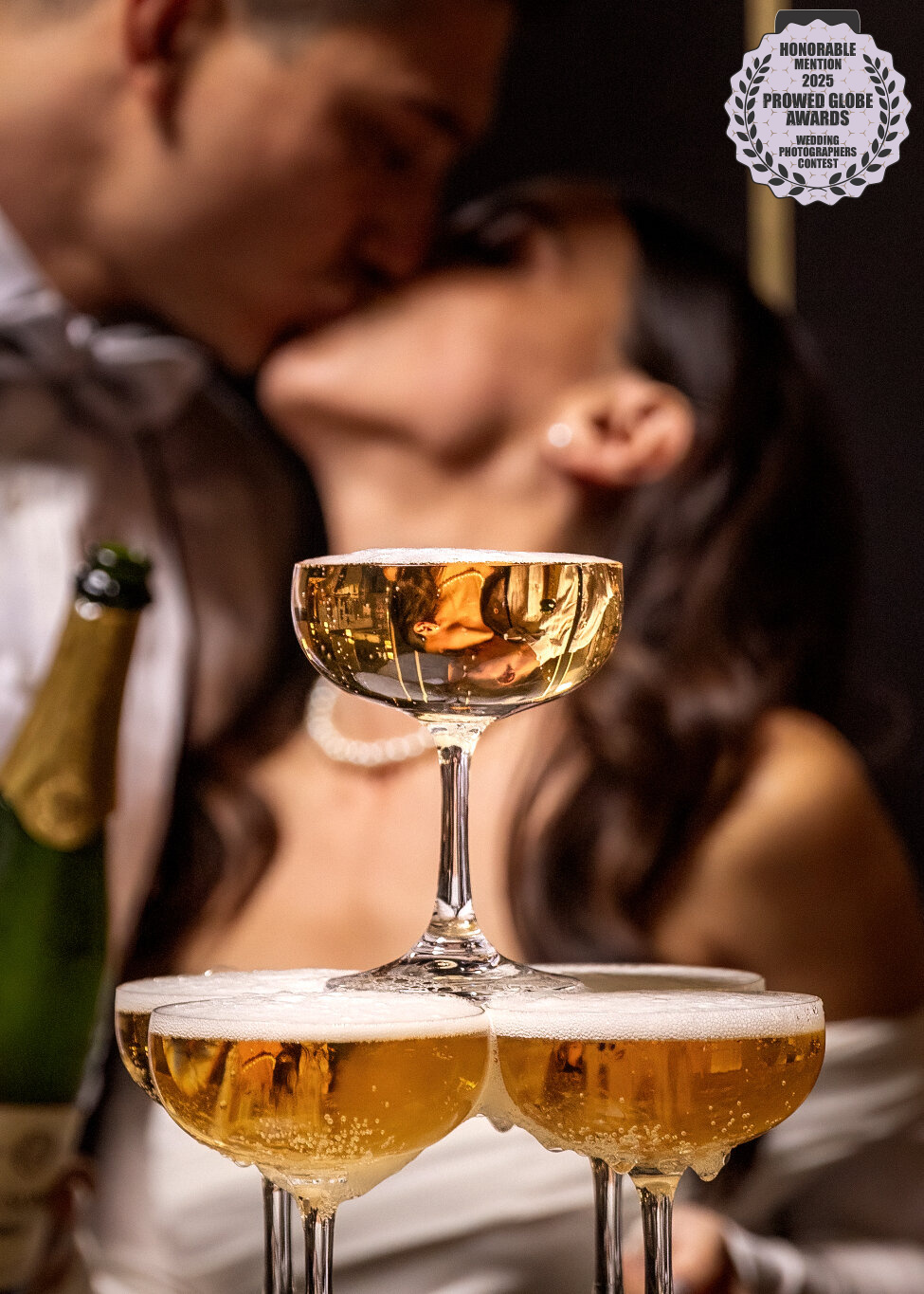2025 ProWed Global Award Honorable Mention Photo of a bride and groom kissing behind a champagne tower with their reflection captured in a coupe glass at the wedding reception at Club Charles in downtown Kerrville, Texas.